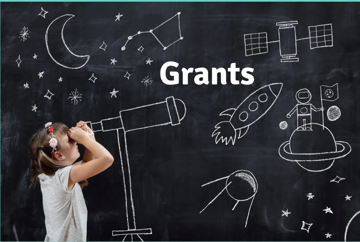 A young girl looks through a pretend chalk telescope drawn on a blackboard filled with space-themed doodles, including stars, a rocket, an astronaut on a planet, and the word ‘Grants’ written in bold white letters