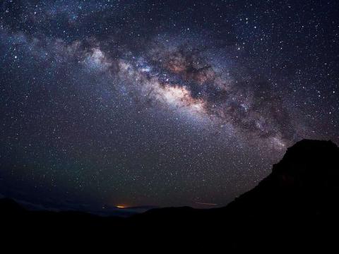 A photograph of the Milky Way galaxy in a dark night sky - it stretches east-west across the photo with the silhouettes of hills seen in the foreground.