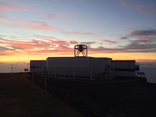 An open dome on a mountaintop with a telescope pointing up at the sky. The sky is blue-lilac with some clouds. The higher clouds are lit up by the sun beneath, making them an orange-red. There are white clouds in shade below the mountaintop.