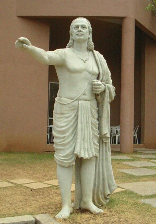 A white statue of a man standing. His arm is raised as he points. He has wavy shoulder-length hair and wear cloth around his waist. A cloth is also draped over his shoulder, gathered in his arm, which falls to the ground.