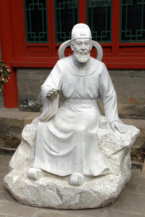 A white sculpture depicting a man sitting on a rock-like seat. He is bearded and wearing robes, and one of his hands is raised.