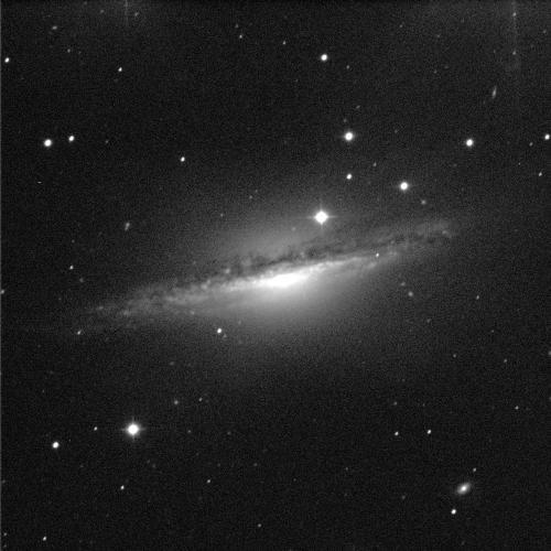 A spiral galaxy, positioned edge-on, against a black background with white dots of stars in various sizes. The centre of the galaxy shines bright white, but it obscured by a band of dust that wraps around it. The band is various shades of grey.