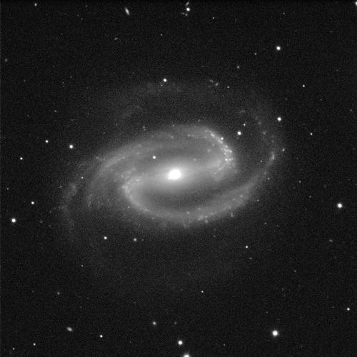 An image of a barred spiral galaxy. It has a brightly glowing white centre. There is a grey fuzzy cloudy line stretching behind the circle, which then extends out at either end to form curling arm-like structures around the galaxy. The background is black with a few dots of white stars.