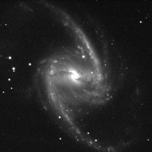 A spiral galaxy angled face-on against a black background with white dots of stars. The galaxy has a small, circular, bright, white centre with a bar-like structure, and two dusty grey spiral arms sweeping upwards and downwards.