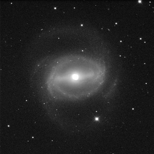 An image of a barred spiral galaxy. It has a brightly glowing white centre. There is a grey fuzzy cloudy line stretching behind the circle, which then extends out at either end to form curling arm-like structures around the galaxy. The background is black with a few dots of white stars.