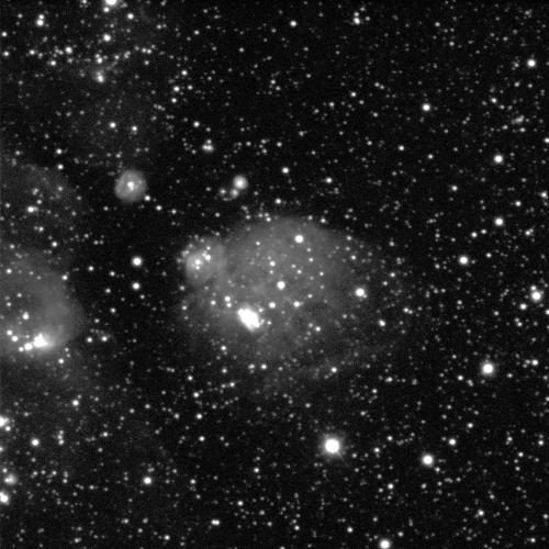 A grey nebula against a black background with white dots of stars in various sizes. The nebula is a small sphere in the centre with another, smaller sphere above it on the right. There is also a patchy, grey, cloudy area on the image right, suggesting the nebula continues in that direction.