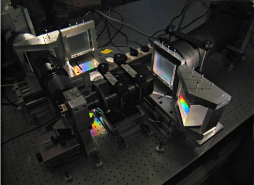 Photograph of a scientific instrument setup featuring optical components and prisms, with light being dispersed into a spectrum, used for astronomical observations or experiments.