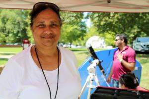 An Indian woman facing the camera and smiling. Her hair is tied back and she wears sunglasses on top of her head. She stands under a canopy-like tent, some of which can be seen running horizontally across the top of the photo. In the background is a sunny field with trees and a man standing beside a telescope.