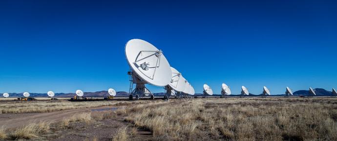Numerous large antenna placed at different distance, all pointing towards the upper right of the image. The sky is bright blue and clear, and some hills can be seen in the far distance.