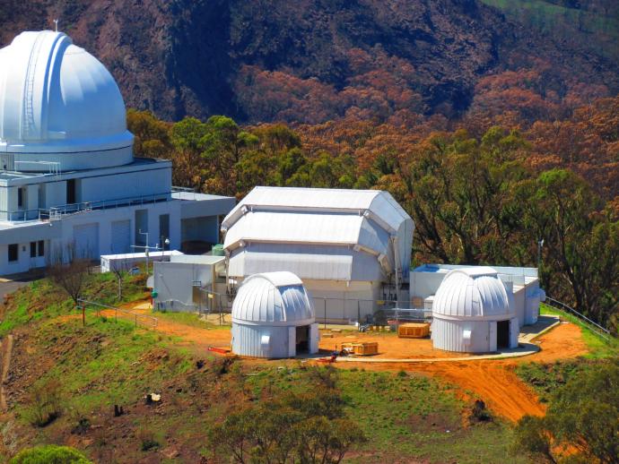White telescope domes of various sizes on a mountaintop. The ground is green and orange, and in the background, there are some trees.