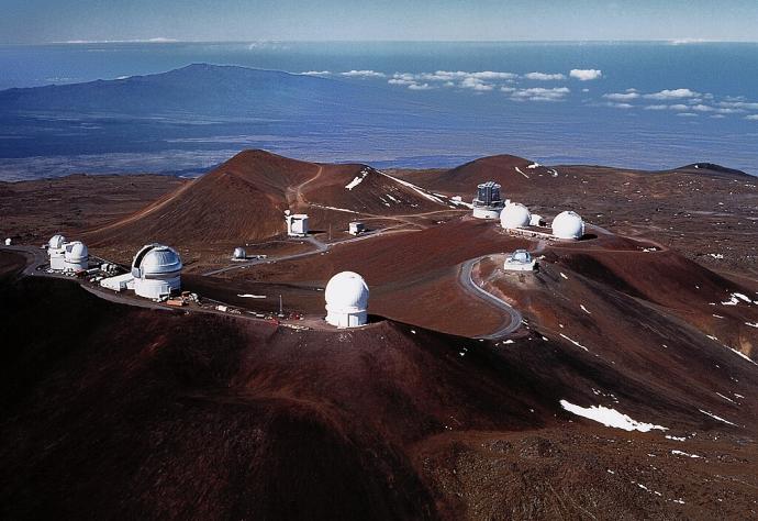 A mountain top with multiple white domes, containing telescopes, placed in different locations. In the background of the image, the landscape below is visible into the distance.