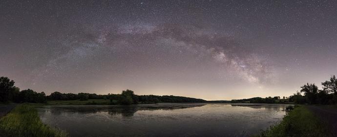 A lake at night with dark silhouettes of trees around the edge. Most of the image is focused on the night sky, where a bright arc sweeps upwards and then down again. Dark brown-red clouds cover sections of the brighter patches in areas. Small, white dots of stars are scattered everywhere in the sky.