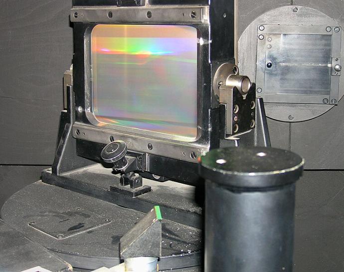 A transparent square held up within a metal frame. There are rainbow colours across the surface of the square.