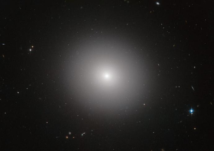 Some small stars and galaxies of a range of colours against a black background. A larger, bright white-yellow sphere is at the centre, and a diffuse glow shines outwards all around it.