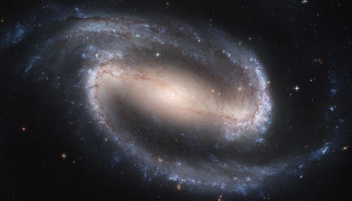 A barred spiral galaxy. There is a bright yellow-orange sphere in the middle with a glow around it. The glow appears like a bar through the centre. From this, blue-grey clouds swirl outwards, forming the spiral arms. Dots of stars are visible in the arms, mostly bright white and blue in colour, but some further out from the centre are more reddish in colour. There are also stars against the black background