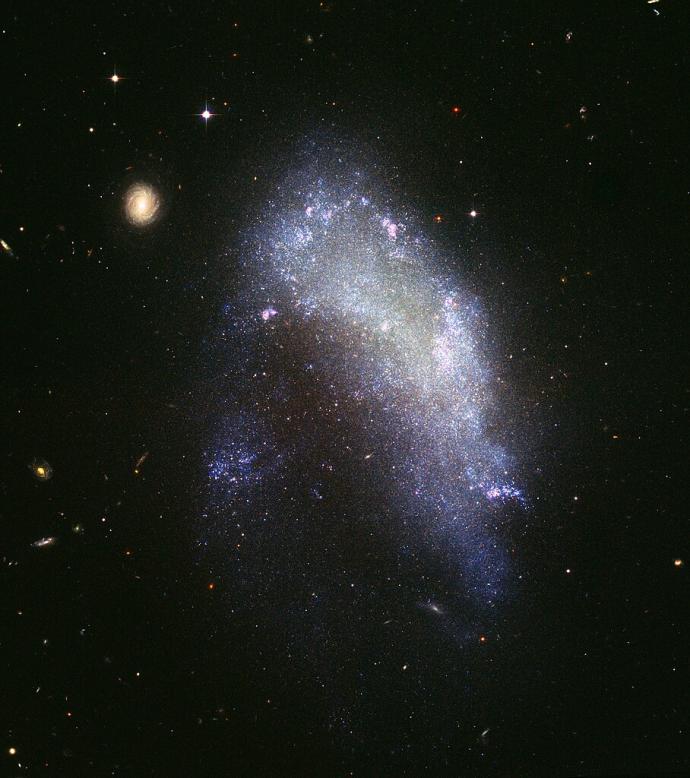 There is a large, cloudy, fuzzy, purple-blue shape in the centre, seemingly like a smudge. Small dots of stars are visible against a black background, varying in size and colour. A few galaxies are seen too, the most notable being towards the upper left, a swirly orange shape.