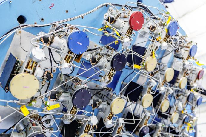 Many small, circles (in colours blue, gold, red, and black) mounted on metal frames to a surface. Thin, silver bars connect these small systems together. The bars run in straight lines that turn in all directions, and go forwards and backwards, snaking between the mounts. There are also wire cables visible.