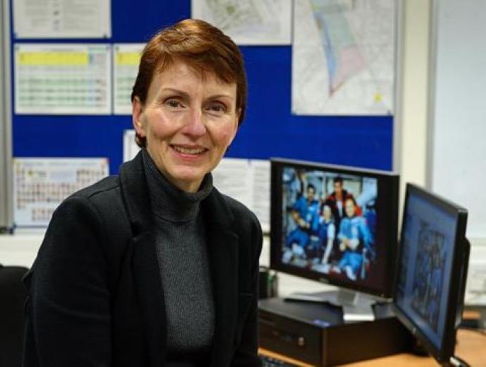 A photograph of Dr. Helen Sharman, the first British astronaut, seated in an office setting. She is smiling and wearing a dark jacket, with a computer monitor in the background displaying a group image of astronauts